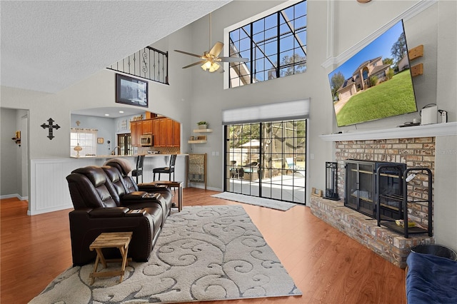 living room with a brick fireplace, ceiling fan, a towering ceiling, a textured ceiling, and light hardwood / wood-style floors