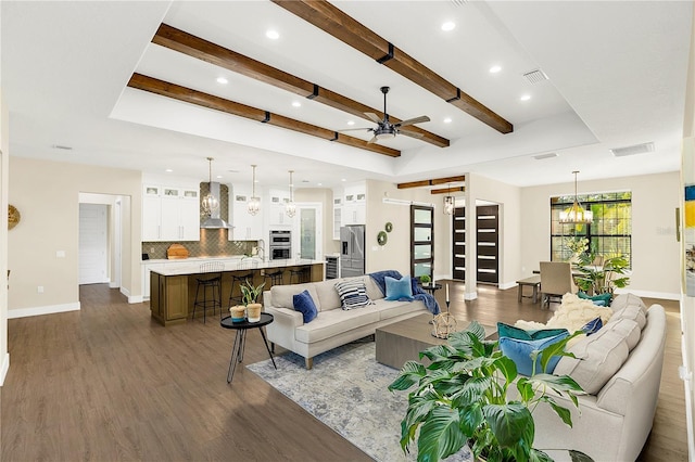 living room with ceiling fan with notable chandelier, beam ceiling, a raised ceiling, and dark wood-type flooring