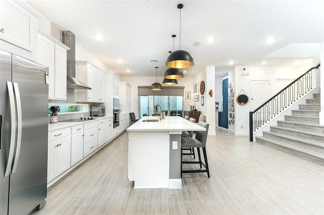 kitchen with white cabinetry, wall chimney exhaust hood, stainless steel fridge with ice dispenser, decorative light fixtures, and a kitchen island with sink