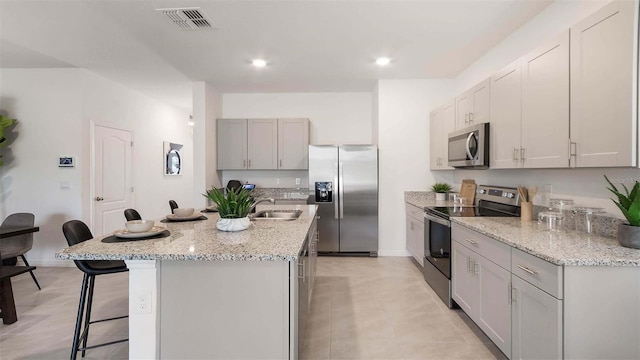 kitchen with light stone countertops, sink, stainless steel appliances, an island with sink, and a kitchen bar