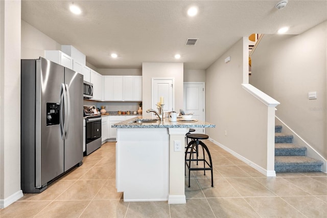 kitchen featuring a kitchen bar, a kitchen island with sink, white cabinets, appliances with stainless steel finishes, and light stone counters