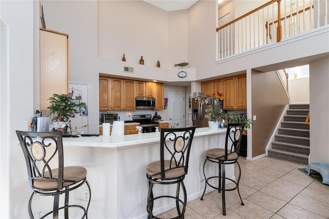 kitchen featuring kitchen peninsula, a towering ceiling, a breakfast bar, light tile patterned flooring, and appliances with stainless steel finishes