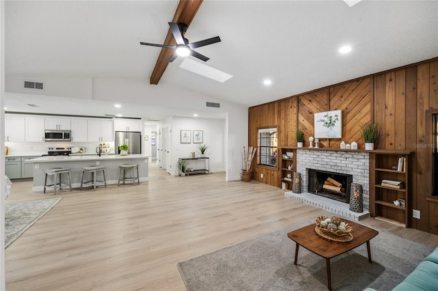 living room with light wood-type flooring, ceiling fan, wooden walls, a fireplace, and vaulted ceiling with skylight