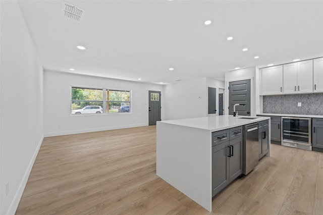 kitchen featuring backsplash, stainless steel dishwasher, gray cabinets, an island with sink, and beverage cooler