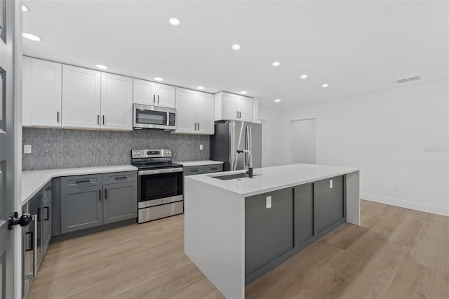 kitchen with gray cabinetry, white cabinets, an island with sink, light hardwood / wood-style floors, and stainless steel appliances