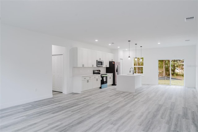 kitchen with appliances with stainless steel finishes, hanging light fixtures, white cabinets, light wood-type flooring, and a kitchen island with sink