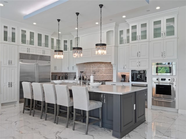 kitchen with white cabinets, hanging light fixtures, built in appliances, an island with sink, and tasteful backsplash