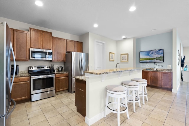 kitchen with stainless steel appliances, light stone counters, a kitchen island with sink, a breakfast bar, and light tile patterned floors