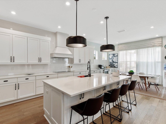 kitchen with black electric stovetop, custom range hood, a kitchen island with sink, sink, and hanging light fixtures