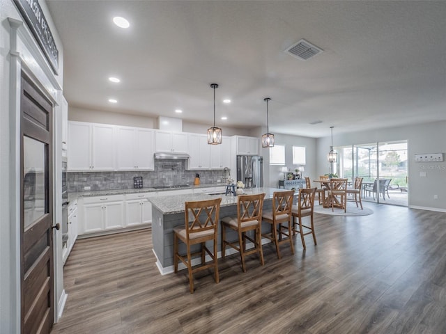 kitchen featuring decorative light fixtures, an island with sink, white cabinets, stainless steel fridge, and light stone counters