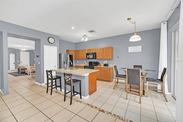 kitchen with light stone counters, a kitchen island with sink, black appliances, light tile patterned floors, and decorative light fixtures