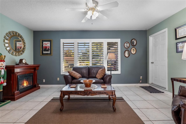 living area featuring baseboards, a glass covered fireplace, ceiling fan, a textured ceiling, and light tile patterned flooring