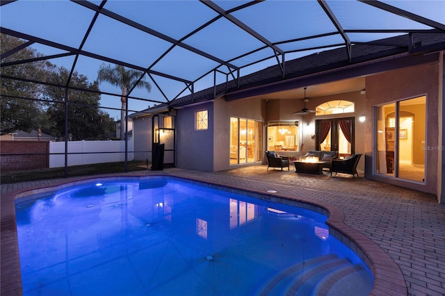 pool at dusk with ceiling fan, a patio area, an outdoor fire pit, and glass enclosure