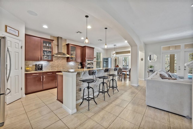 kitchen featuring stainless steel refrigerator, tasteful backsplash, an island with sink, decorative light fixtures, and wall chimney exhaust hood