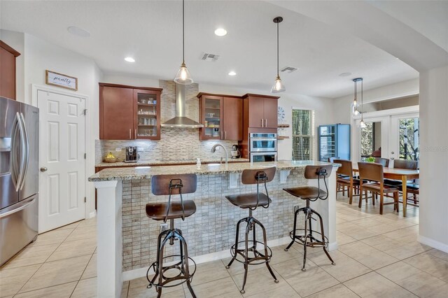 kitchen with appliances with stainless steel finishes, light stone counters, a center island with sink, decorative light fixtures, and wall chimney exhaust hood