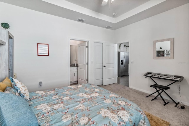 carpeted bedroom with a tray ceiling, connected bathroom, ceiling fan, and stainless steel refrigerator with ice dispenser