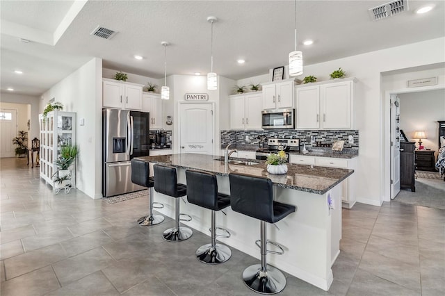 kitchen with dark stone counters, a center island with sink, white cabinets, appliances with stainless steel finishes, and decorative light fixtures