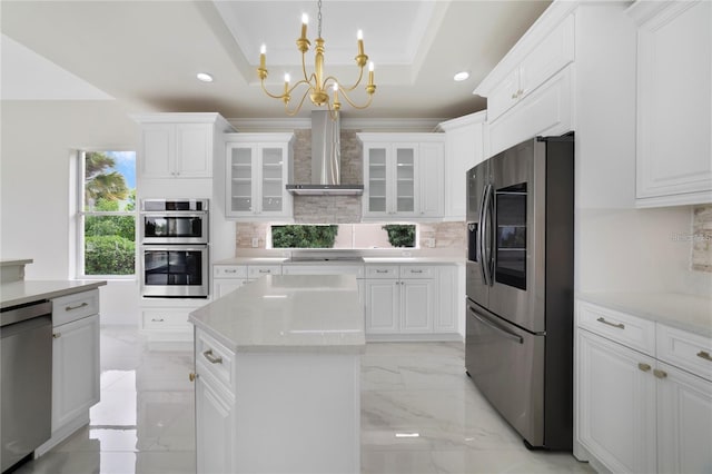 kitchen with an inviting chandelier, white cabinets, wall chimney range hood, a tray ceiling, and stainless steel appliances