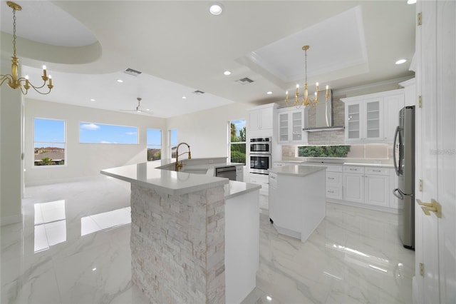 kitchen featuring stainless steel appliances, hanging light fixtures, wall chimney range hood, a kitchen island with sink, and white cabinets