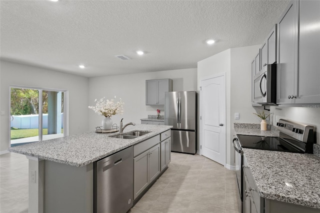 kitchen with a kitchen island with sink, gray cabinets, appliances with stainless steel finishes, light stone counters, and sink