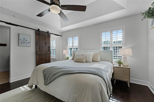 bedroom with ceiling fan, a barn door, a tray ceiling, multiple windows, and dark hardwood / wood-style flooring