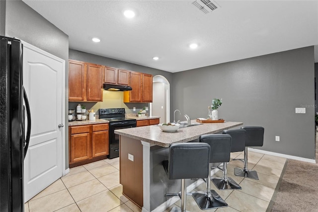 kitchen with sink, light tile patterned floors, an island with sink, black appliances, and a breakfast bar