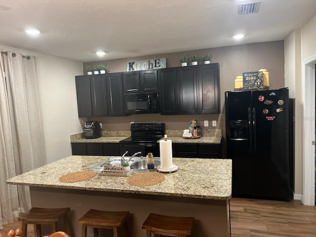 kitchen featuring a center island with sink, a kitchen bar, black appliances, and light stone countertops