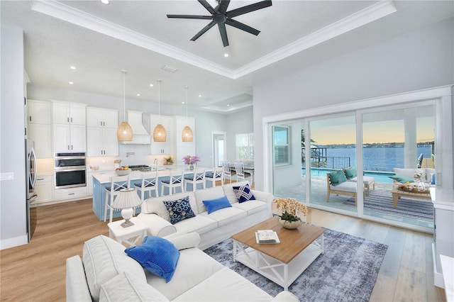 living room with ceiling fan, crown molding, a tray ceiling, a water view, and light wood-type flooring