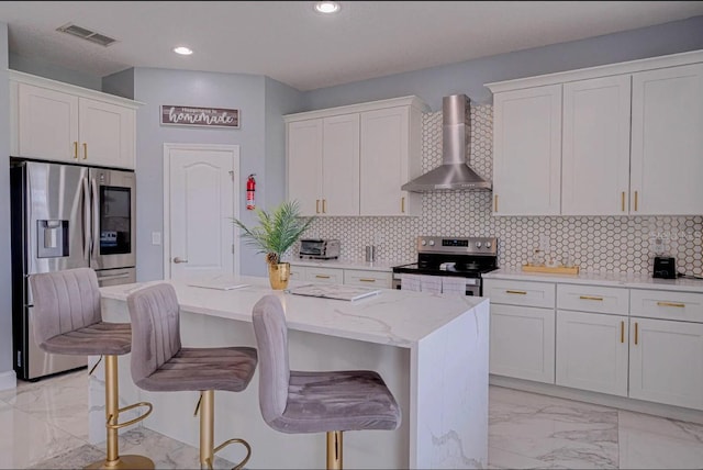 kitchen with wall chimney range hood, a kitchen island, white cabinetry, stainless steel appliances, and light stone counters