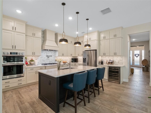 kitchen featuring appliances with stainless steel finishes, wine cooler, custom exhaust hood, hanging light fixtures, and a center island with sink