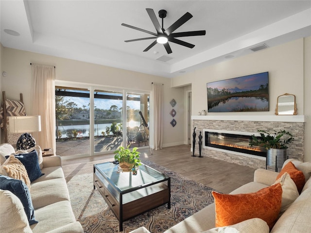 living room featuring a stone fireplace, light hardwood / wood-style floors, a raised ceiling, and ceiling fan