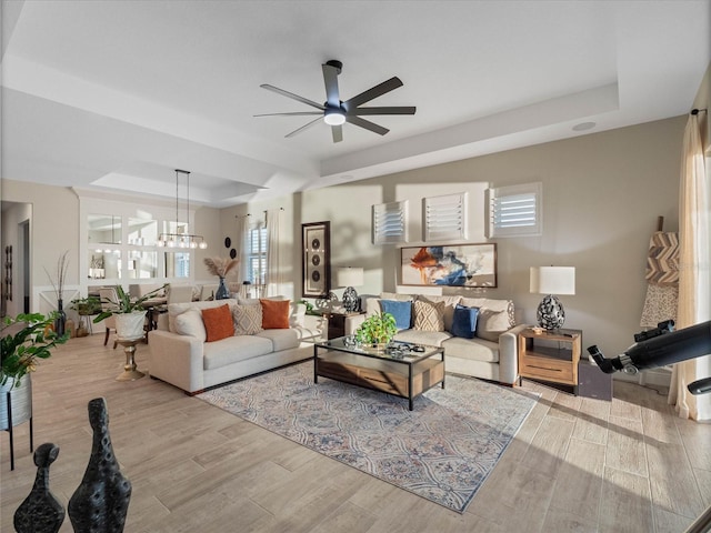 living room with ceiling fan with notable chandelier, a tray ceiling, and light hardwood / wood-style floors