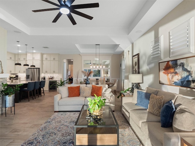 living room featuring light hardwood / wood-style floors, ceiling fan with notable chandelier, beverage cooler, and a raised ceiling