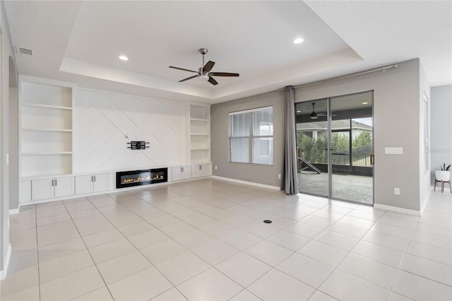 unfurnished living room featuring baseboards, built in features, a glass covered fireplace, a tray ceiling, and light tile patterned flooring