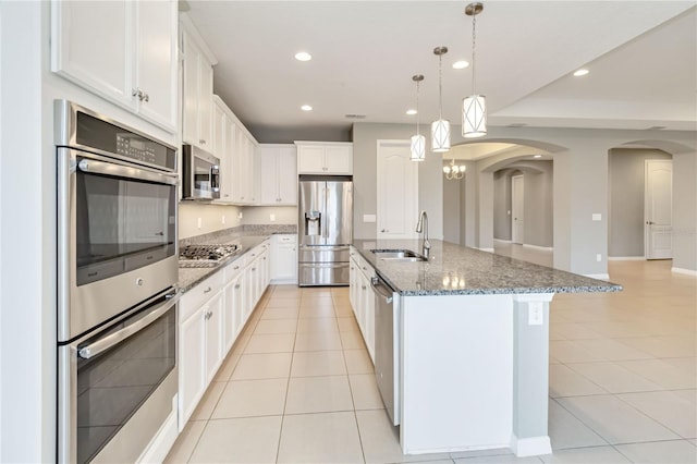 kitchen featuring arched walkways, dark stone counters, appliances with stainless steel finishes, a sink, and light tile patterned flooring