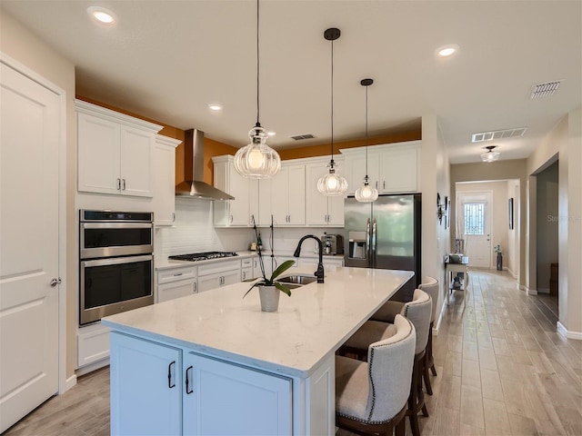 kitchen featuring wall chimney range hood, an island with sink, white cabinets, and appliances with stainless steel finishes