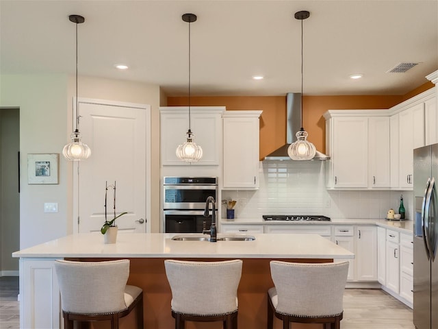 kitchen with a center island with sink, appliances with stainless steel finishes, wall chimney range hood, and white cabinets