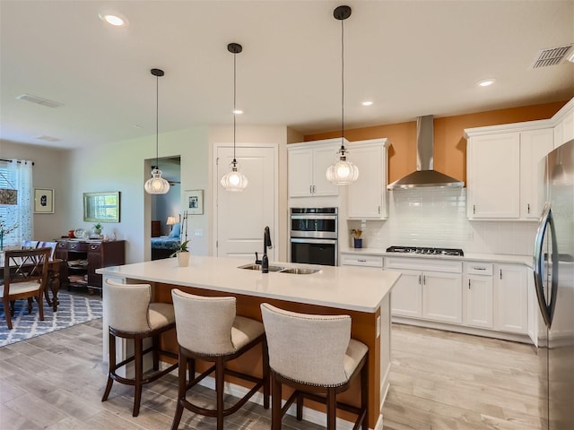 kitchen featuring white cabinetry, stainless steel appliances, an island with sink, decorative light fixtures, and wall chimney exhaust hood