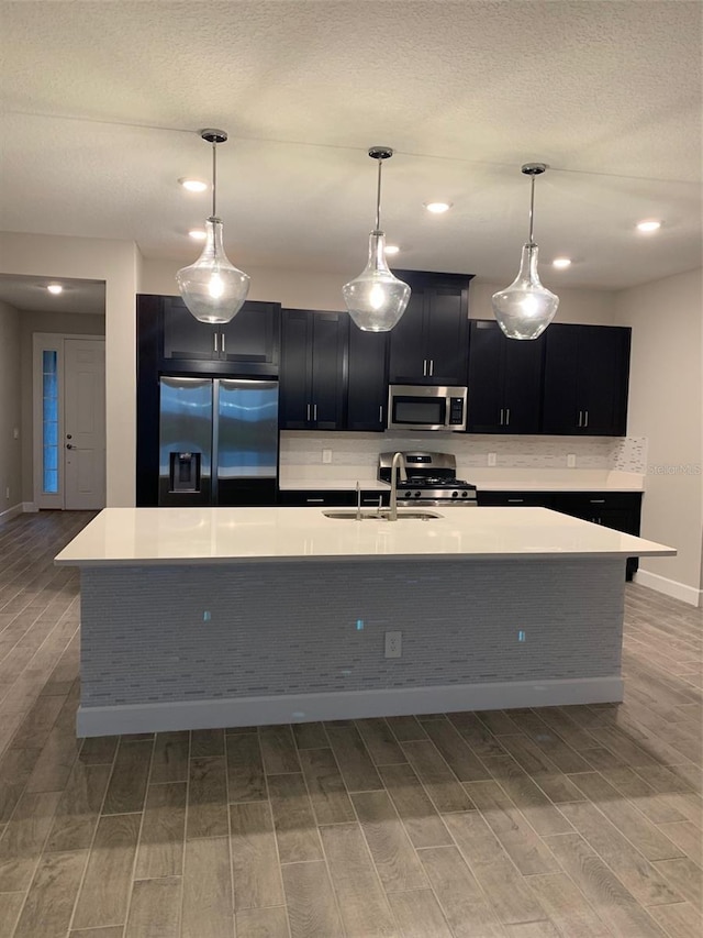 kitchen featuring a textured ceiling, pendant lighting, stainless steel appliances, a kitchen island with sink, and sink