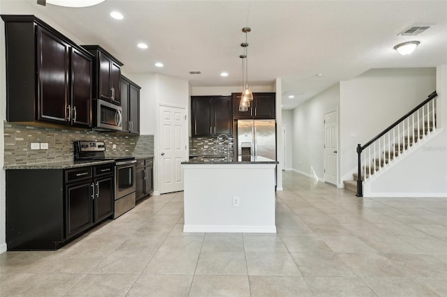 kitchen featuring a kitchen island with sink, dark stone counters, decorative backsplash, appliances with stainless steel finishes, and decorative light fixtures