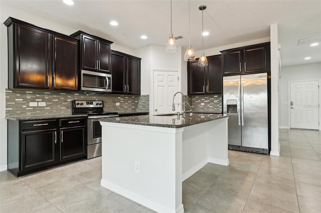 kitchen with appliances with stainless steel finishes, a kitchen island with sink, sink, dark stone countertops, and hanging light fixtures