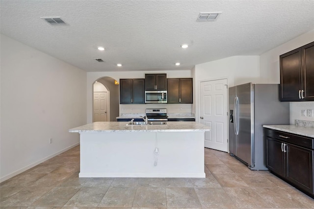 kitchen featuring a textured ceiling, a center island with sink, dark brown cabinets, and appliances with stainless steel finishes