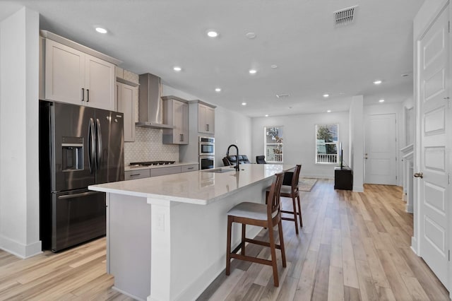 kitchen with stainless steel appliances, wall chimney exhaust hood, light hardwood / wood-style floors, a center island with sink, and a breakfast bar