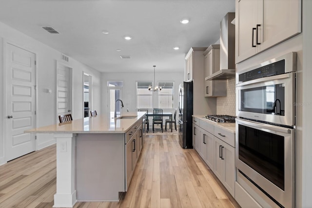 kitchen featuring wall chimney exhaust hood, hanging light fixtures, an island with sink, gray cabinetry, and appliances with stainless steel finishes