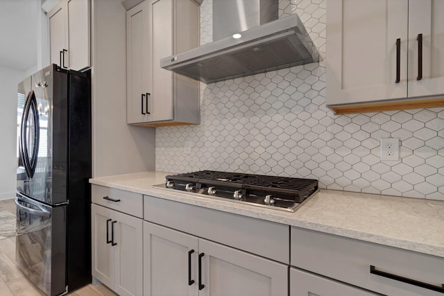 kitchen with light stone countertops, wall chimney range hood, tasteful backsplash, black refrigerator, and stainless steel gas cooktop