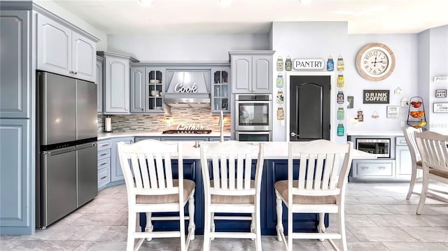 kitchen with gray cabinets, premium range hood, stainless steel appliances, a kitchen breakfast bar, and a center island with sink