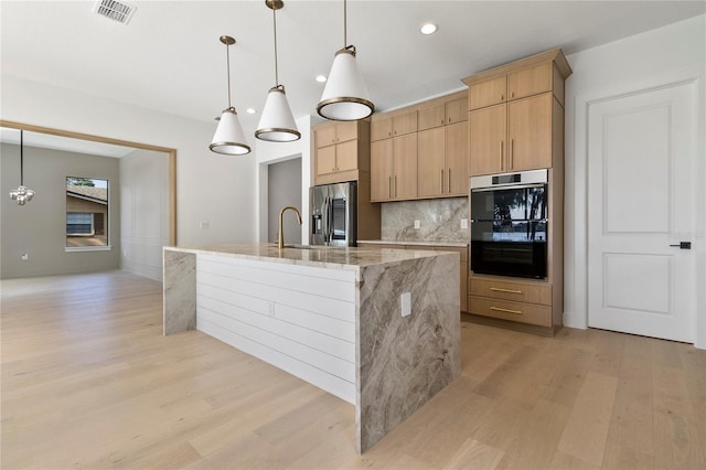 kitchen featuring light brown cabinets, a kitchen island with sink, hanging light fixtures, light stone countertops, and double oven