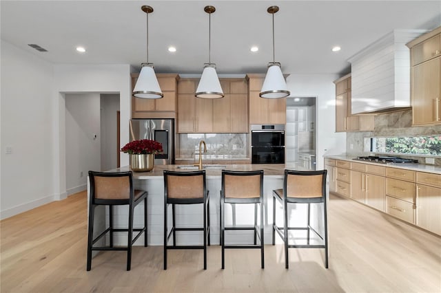 kitchen with a center island with sink, appliances with stainless steel finishes, light brown cabinetry, and decorative light fixtures