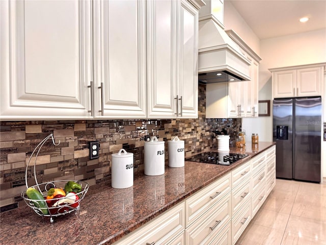 kitchen with custom range hood, black electric stovetop, stainless steel fridge with ice dispenser, light tile patterned floors, and dark stone countertops