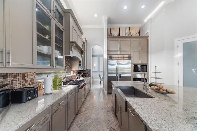 kitchen featuring sink, built in appliances, decorative backsplash, ornamental molding, and light stone counters